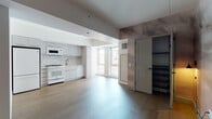 This single-floor residence offers a thoughtfully designed open-plan layout that seamlessly integrates the kitchen and living areas. The kitchen is a standout feature, equipped with white appliances including a gas range, built-in microwave, refrigerator, and dishwasher. Gray cabinetry with brushed gold hardware complements a striking marble-style backsplash, while the countertops maintain a clean, minimal aesthetic. A gold-toned faucet adds a refined detail to the sink area.

Light hardwood flooring runs throughout the main living spaces, lending warmth and visual continuity. Large windows allow natural light to fill the interior, offering views of the surrounding urban environment. A dramatic artistic accent wall, featuring a soft, atmospheric mural in muted pink and gray tones, creates a bold focal point within the living area. Built-in shelving and a closet with paneled doors provide practical storage solutions.

The bathroom is finished in a darker palette, with deep charcoal subway-tiled walls surrounding a soaking tub and shower combination. Slate-style floor tiles, a louvered closet door, and a wood-toned vanity complete the sophisticated look. The overall ambiance is modern yet warm, blending contemporary finishes with artistic touches throughout.