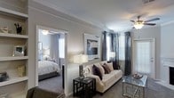 This single-floor residence offers a cohesive and elegantly appointed interior designed for comfortable living. The living area serves as the heart of the space, featuring plush carpeting, crown molding, and a ceiling fan that adds both function and a classic architectural touch. Large windows dressed with layered blinds and floor-length drapes in muted tones allow natural light to fill the room while maintaining privacy. The living area is spacious enough to accommodate a full sofa and accent furniture, creating a welcoming atmosphere.

A wide open doorway connects the living area to the bedroom, which mirrors the same refined design language — complete with its own ceiling fan, large windows, and soft carpeting. The bedroom comfortably fits a full-sized bed and additional furnishings, maintaining a calm and restful ambiance consistent with the rest of the home.

Throughout the space, neutral wall tones in warm gray provide a versatile backdrop that complements the decorative accents. The consistent use of crown molding across both rooms lends the interior a polished, cohesive feel. Overall, the space balances practical livability with understated elegance, making it a comfortable and visually appealing single-floor residence.