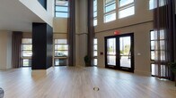 This expansive lobby and amenity space makes an immediate impression with its dramatic double-height ceilings and floor-to-ceiling windows that flood the interior with natural light. The main entry is framed by oversized black-framed glass doors flanked by tall curtain panels, leading into a grand open floor plan finished with wide-plank light wood flooring throughout. A standout feature is the illuminated brick accent wall interlaced with bold diagonal black beams, serving as a dramatic focal point for one of several curated seating areas. That lounge is anchored by a curved sectional sofa, sculptural accent chairs, and a round area rug, creating an intimate gathering space within the larger volume. Deeper into the floor, a sleek reception desk with blue upholstered seating offers a more formal service area, set against a backdrop of warm vertical slat partitions that define zones without closing off the open-plan layout. A mezzanine level with open-rail balustrades overlooks the main floor, adding vertical depth and architectural interest. Pendant globe chandeliers, recessed lighting, and curated contemporary artwork — including vibrant yellow abstract pieces — contribute to a polished, modern aesthetic. Potted plants and thoughtfully arranged furnishings complete the welcoming, design-forward atmosphere.