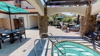 Located in Stevenson Ranch, CA, this outdoor amenity area offers a well-appointed resort-style experience defined by thoughtful design and a variety of recreational features. The centerpiece is a generously sized swimming pool with a smooth concrete surround, lined with lounge chairs and framed by towering palm trees and lush greenery against a backdrop of rolling hills. Adjacent to the pool, a covered spa provides a relaxing retreat, sheltered beneath a shade structure supported by distinctive stacked-stone columns that lend a warm, Mediterranean character to the space. The expansive paver patio serves as a social hub, featuring multiple seating zones including a sectional lounge surrounding a stone fire pit, ideal for evening gatherings. Umbrella-shaded dining tables offer casual al fresco options throughout the day. A built-in outdoor kitchen with a stone surround adds functional convenience for entertaining. Wrought iron fencing defines the perimeter while maintaining open sightlines to the surrounding landscape, including mature jacaranda, pine, and palm trees. The Spanish-influenced architecture, with its clay tile rooflines and stucco exteriors, ties the entire outdoor environment together into a cohesive and inviting community amenity space.