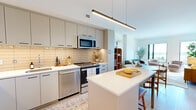 Located at 1796 Chain Bridge Rd in McLean, VA, this thoughtfully designed one-bedroom residence offers a well-appointed single-floor layout that maximizes both function and style. The open-concept kitchen and living area forms the heart of the home, where sleek flat-front cabinetry in a soft grey tone pairs with a warm tile backsplash, quartz countertops, and a full suite of stainless steel appliances. A central island with bar seating provides additional workspace and a natural gathering point. Light hardwood flooring runs throughout, lending warmth and continuity to every room. The living room is bathed in natural light from large floor-to-ceiling windows that frame sweeping views of the McLean skyline and surrounding greenery. A private balcony extends the living space outdoors, offering an elevated vantage point of the landscape below. The bedroom features a distinctive accent wall with geometric wallcovering, large windows with balcony access, and ample room for a full furniture arrangement. A dedicated walk-in closet with adjustable shelving and hanging systems provides generous storage. Clean white walls, modern fixtures, and a cohesive neutral palette give the entire residence a bright, contemporary ambiance throughout.