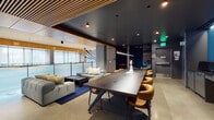 Located at 1150 Clay St in Oakland, CA, this single-floor amenity space delivers a sophisticated, design-forward environment that blends collaborative functionality with refined aesthetics. The open-plan layout is anchored by a substantial dark wood communal table surrounded by curved cognac leather chairs, making it well-suited for group work, meetings, or casual gatherings. Adjacent to the table, a generous lounge area features plush sectional sofas arranged on a deep navy area rug, providing a comfortable setting for relaxed conversation or focused downtime. Exposed concrete columns and polished concrete floors lend an industrial character, while slatted wood ceiling panels and warm accent lighting introduce texture and warmth throughout. Glass partitions along one wall allow natural light to filter into the space, maintaining an open and airy atmosphere. Toward the rear, a pair of intimate private pods enclosed in dark-toned glazing offer quiet retreat for focused work or small meetings. A sleek kitchenette rounds out the space, showcasing striking black marble countertops with gold veining, a brass undermount sink, and bold teal cabinetry. Decorative artwork and sculptural accents further elevate the overall ambiance, resulting in a polished, contemporary space designed for both productivity and comfort.