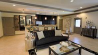 Located at 200-A Heron Cove Dr in Enterprise, Alabama, this single-story space presents a refined and sophisticated interior defined by an open-concept layout that seamlessly connects the living and kitchen areas. The living area is anchored by a tufted black velvet sofa accented with geometric throw pillows, complemented by sleek metallic accent tables and a glass-top coffee table that reflect the space's glamorous aesthetic. Decorative touches such as sunflower arrangements, ornate vases, and statement wall art add warmth and personality throughout.

The kitchen is a standout feature, showcasing rich, dark espresso cabinetry paired with light countertops and stainless steel appliances. Pendant lighting with warm amber tones hangs above the kitchen bar, casting an inviting glow over the space. The bar-height counter with metallic paneling serves as a natural divider between the kitchen and living areas, offering both functionality and visual elegance.

Light-toned tile flooring runs throughout, enhancing the sense of openness and providing a neutral foundation that allows the bold furnishings and dark cabinetry to take center stage. Recessed lighting and a tray ceiling detail in the living area add architectural depth, contributing to the overall polished and upscale ambiance of this well-appointed space.