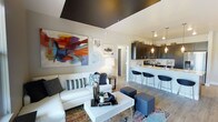 This single-floor space showcases a thoughtfully designed open-concept layout that combines the living and kitchen areas into one cohesive environment. The living area is anchored by a white sectional sofa accented with patterned throw pillows, paired with a clean-lined white coffee table and navy ottoman seating. A striking large-scale abstract canvas in warm reds, blues, and oranges adds a vibrant focal point to the wall, complemented by a curated gallery arrangement alongside it. One of the most distinctive architectural features is the bold dark-painted tray ceiling above the living area, creating a dramatic contrast against the lighter surrounding walls and adding a sense of defined depth to the space. Light wood-toned flooring runs throughout, contributing warmth and continuity. The kitchen is finished with dark cabinetry, stainless steel appliances, and a light-colored countertop that extends into a breakfast bar with sleek modern bar stools. Pendant lights hang above the bar, providing focused ambient lighting. The layout flows efficiently, with a hallway visible in the background suggesting additional rooms beyond the main living space.