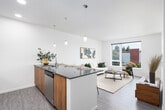 This thoughtfully designed single-floor residence blends modern aesthetics with functional living. The open-concept layout seamlessly connects the kitchen, dining, and living areas, creating a cohesive and airy atmosphere. The kitchen is a standout feature, showcasing dark quartz countertops, a spacious breakfast bar with seating for multiple guests, stainless steel appliances including a refrigerator, microwave, and range, and a combination of warm wood-toned and dark cabinetry accented by a tile backsplash. Stylish pendant lighting illuminates the kitchen and dining area, adding an inviting warmth. The living room benefits from a large picture window that draws in abundant natural light and offers views of the surrounding neighborhood, complementing the wood-style flooring that runs throughout the space. The bedroom is a calm retreat with sliding panel closet doors offering generous storage and a ceiling fan for comfort. A second, smaller bedroom or flex room is also present, providing additional versatility. The bathroom is clean and well-appointed, featuring a soaking tub with shower combination, wood-toned vanity with a white countertop, and wood-look tile flooring. The unit's crisp white walls and consistent modern finishes create a cohesive and contemporary living environment throughout.