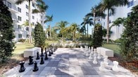This inviting outdoor space showcases the hallmarks of a thoughtfully designed resort-style amenity area. At its centerpiece is a large-format chess board laid in crisp, light-toned pavers, complete with oversized black and white chess pieces that create an engaging, interactive focal point for residents and guests alike. The surrounding landscape is richly appointed with towering palm trees, manicured hedges, and lush tropical greenery that frame the space and lend a sense of privacy and serenity. Elegant post lanterns line the area, providing both a refined aesthetic and practical illumination for evening enjoyment. Beyond the chess court, the grounds open up to reveal well-maintained lawn areas and additional landscaped features, suggesting a broader outdoor campus with multiple leisure amenities. The modern white-facade buildings flanking the space reinforce a clean, contemporary architectural character throughout the property. Abundant natural light and open blue skies create a bright, welcoming atmosphere, making this outdoor destination ideal for relaxed social gatherings and leisurely recreation in a tropical setting.