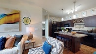 This 2-bedroom, 2-bathroom residence is thoughtfully designed to maximize comfort and functionality across a single floor. The open-concept living and kitchen area creates a seamless flow, making the space feel welcoming and connected. The kitchen is a standout feature, showcasing rich dark cabinetry, granite countertops, and a breakfast bar with bar seating — ideal for casual dining or entertaining. Pendant lighting and track lighting illuminate the space, complementing the warm hardwood-style flooring that runs throughout. Black appliances add a sleek, modern contrast to the cabinetry. The living area is bright and inviting, with neutral wall tones that provide a versatile backdrop for a variety of design styles. A glimpse into the hallway reveals a well-lit bathroom with classic finishes, reflecting the consistent attention to detail found throughout the residence. The layout offers a natural separation between living and sleeping areas, providing privacy without sacrificing openness. Overall, this residence combines contemporary design elements with a practical floor plan, delivering a comfortable and stylish living environment suited for a range of lifestyles.