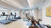 Located at 118 N Prince St in Lancaster, PA, this fitness center offers a thoughtfully designed, single-floor exercise facility divided into three interconnected exercise rooms. The space features light-toned luxury vinyl flooring throughout, crisp white walls, and a clean, modern aesthetic that creates an open and motivating environment.

The main cardio area is bathed in natural light from large arched windows and is equipped with multiple treadmills, a stationary bike, and a stair climber. Linear LED lighting runs along the ceiling, complementing the bright, airy atmosphere. Adjacent to this room, a dedicated cycling studio houses Peloton bikes positioned on individual mats, accompanied by wall-mounted screens for immersive ride experiences. A vibrant piece of cycling-themed artwork adds a playful touch to the space.

A separate free weights and functional training area provides a well-organized rack of dumbbells ranging in various sizes, an adjustable workout bench, and a multi-use vertical training rig. A centrally located accessory station offers resistance bands, kettlebells, stability balls, and other small equipment. Colorful artwork is displayed throughout, contributing to an energetic and welcoming atmosphere. The facility also includes accessible amenities and clearly marked exits, ensuring a safe and comfortable experience for all users.