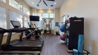 This single-level fitness center is designed to support a comprehensive workout experience, combining thoughtful architecture with a strong selection of modern equipment. The space features soaring ceilings that lend an open, airy feel, while multiple large windows fitted with plantation shutters allow abundant natural light to fill the room. Warm-toned wood-look flooring runs throughout, complementing the neutral, light-colored walls and recessed lighting overhead.

The cardio zone is anchored by multiple Life Fitness treadmills equipped with interactive touchscreen displays, alongside an elliptical and stationary bike. A wall-mounted flat-screen television provides additional entertainment during workouts. At the center of the room, a multi-functional cable cross-trainer serves as the primary strength training station, suitable for a wide range of resistance exercises.

Along one wall, a dedicated storage rack holds an assortment of kettlebells, medicine balls, and stability balls, offering versatile options for functional training. TRX suspension straps are mounted to another wall, enabling bodyweight-based training. A standout feature is the interactive smart mirror, which provides guided workout programming directly from the floor.

The fitness center is well-organized, with posted fitness center rules and a clean, professional atmosphere that encourages regular use.