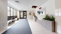 Located at 25 Parkway Forest Dr in Toronto's Parkway Forest neighbourhood, this building lobby serves as the primary entrance and reception area for residents and visitors. The space is thoughtfully laid out across a single level, offering a comfortable and functional welcome area.

Upon entry, a broad runner carpet guides visitors through the main corridor toward a set of stainless steel elevators, framed by distinctive travertine-style stone wall panels that lend a timeless, refined character to the space. The flooring transitions between large-format cream-coloured tiles and grey carpet, clearly delineating pathways and waiting zones.

A built-in reception desk with a wall-mounted mirror and decorative planters filled with lush greenery anchors one side of the lobby, adding a fresh, welcoming touch. Adjacent to the elevators, a sleek, sculptural bench provides a modern seating option for waiting residents or guests. Large windows along one wall invite ample natural light, brightening the interior and providing views of the surrounding exterior.

Overhead, a geometric pendant light fixture with warm-toned accents adds a decorative focal point near the entrance. The lobby also features a bulletin board area and informational displays, supporting building communications. The overall ambiance is clean, practical, and well-maintained, reflecting a community-focused residential environment.
