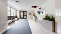 Located at 25 Parkway Forest Dr in Toronto's Parkway Forest neighbourhood, this building lobby serves as the primary entrance and reception area for residents and visitors. The space is thoughtfully laid out across a single level, offering a comfortable and functional welcome area.

Upon entry, a broad runner carpet guides visitors through the main corridor toward a set of stainless steel elevators, framed by distinctive travertine-style stone wall panels that lend a timeless, refined character to the space. The flooring transitions between large-format cream-coloured tiles and grey carpet, clearly delineating pathways and waiting zones.

A built-in reception desk with a wall-mounted mirror and decorative planters filled with lush greenery anchors one side of the lobby, adding a fresh, welcoming touch. Adjacent to the elevators, a sleek, sculptural bench provides a modern seating option for waiting residents or guests. Large windows along one wall invite ample natural light, brightening the interior and providing views of the surrounding exterior.

Overhead, a geometric pendant light fixture with warm-toned accents adds a decorative focal point near the entrance. The lobby also features a bulletin board area and informational displays, supporting building communications. The overall ambiance is clean, practical, and well-maintained, reflecting a community-focused residential environment.