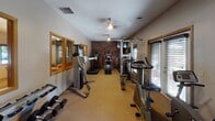 Located at 11360 Robinson Dr NW in Coon Rapids, MN, this single-floor fitness center is a well-equipped exercise facility designed to support a range of workout routines. The space features a distinctive exposed brick accent wall that adds a warm, classic character to the room, complemented by neutral-toned walls and carpet flooring throughout.

The facility is outfitted with a comprehensive selection of cardio equipment, including treadmills, elliptical trainers, a stair climber, and stationary bikes, arranged to allow comfortable movement between machines. A dedicated free weight area includes a multi-tiered dumbbell rack with a full range of weights, alongside a step platform for aerobic and functional training. A cable and strength machine rounds out the equipment offerings, providing users with options for full-body resistance training.

Floor-to-ceiling mirrors line one wall, visually expanding the space and allowing users to monitor their form during workouts. Natural light filters in through slatted blinds on the windows, while ceiling fans provide overhead ventilation and comfort. A pass-through window connects the fitness room to an adjacent lounge area, visible in several views, adding a sense of connectivity within the broader facility. The overall atmosphere is practical and functional, with a layout that efficiently utilizes the available space.
