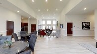 This single-floor space is defined by its open-concept layout and impressive soaring ceilings that create an expansive, airy atmosphere throughout. Light hardwood flooring runs continuously across the entire space, enhancing the sense of openness and providing a clean, neutral foundation for the interior design.

The dining area, positioned toward the front left, features a round dark-topped table accompanied by tufted charcoal upholstered chairs, offering a comfortable and stylish setting. Adjacent to this area, a partial kitchen is visible with dark wood cabinetry and stainless steel appliances, including a refrigerator and microwave.

Further into the space, a well-appointed lounge area is arranged around a central coffee table, featuring a combination of a dark sofa with red accent chairs and a patterned accent chair. A wall-mounted flat-screen television anchors this gathering space.

Natural light floods the interior through a bank of tall windows and glass-paneled doors at the far end, framing views of the outdoors. Recessed lighting is distributed throughout the ceiling, ensuring consistent and even illumination. The overall ambiance is bright, welcoming, and functionally versatile.