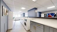 This shared laundry facility offers a spacious, efficiently organized layout designed to accommodate multiple users simultaneously. The room features a clean, modern aesthetic defined by crisp white cabinetry, blue accent walls, and a drop-panel ceiling with recessed fluorescent lighting that keeps the space bright and well-illuminated throughout. A row of commercial-grade front-loading washers and top-loading machines lines the central area, complemented by a matching set of dryers, all equipped with digital control panels for ease of use. Dedicated folding counters with stylish gray cabinetry and gold hardware provide ample workspace for sorting and folding laundry. Vending units stocked with laundry supplies are conveniently positioned along one wall, ensuring essentials are readily available. Natural light filters in through windows along one side of the room, enhancing the open and airy feel of the space. The classic small-tile flooring is durable and easy to maintain, suited to the demands of a high-traffic environment. A utility sink, waste bins, a fire extinguisher, and posted laundry room guidelines round out the practical amenities, making this a fully self-contained and user-friendly facility.
