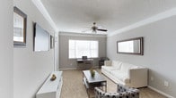 This single-story living space offers a well-appointed interior designed with a neutral color palette of soft grays and whites, creating a calm and inviting atmosphere throughout. The main living area is generously proportioned, featuring plush wall-to-wall carpeting that adds warmth underfoot. Crown molding runs along the ceiling perimeter, lending a polished, refined character to the room.

A ceiling fan with a light fixture is centrally positioned, providing both airflow and ambient lighting. Multiple windows fitted with horizontal blinds allow natural light to filter into the space while maintaining privacy. The walls are painted in a soft gray tone, complementing the white trim and baseboards for a cohesive, modern aesthetic.

The living area is thoughtfully arranged to accommodate both relaxation and productivity, with space for seating and a dedicated desk area positioned near the window. A wall-mounted television area and low-profile media console contribute to the functional layout. The textured ceiling adds subtle visual interest, completing this clean, move-in-ready space.