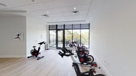 This purpose-built spin studio offers a clean, modern environment designed specifically for indoor cycling workouts. The room is finished with light hardwood-style flooring that lends warmth to the otherwise crisp, white interior, creating a bright and motivating atmosphere for exercise. Multiple stationary cycling bikes are arranged throughout the space, providing ample room for group spin sessions while allowing comfortable spacing between each station. The studio is illuminated by a combination of recessed ceiling lighting and abundant natural light streaming in through large, floor-to-ceiling windows fitted with dark-framed horizontal blinds for adjustable light control. These expansive windows open to an outdoor balcony, offering views of surrounding trees and greenery that add a refreshing, open-air feel to the workout environment. The drop-panel ceiling is fitted with ventilation to ensure proper air circulation during high-intensity sessions. Along one wall, a set of double doors provides additional access points, keeping the flow of the space practical and functional. Motivational fitness artwork adorns the walls, reinforcing the studio's dedicated athletic purpose. The overall design balances simplicity with functionality, making this spin studio a well-equipped and inviting space for cycling enthusiasts of all levels.