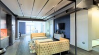 This single-floor space showcases a compelling blend of industrial and contemporary design. The open layout is defined by exposed ceiling ductwork and structural steel beams, which contrast elegantly with the clean white ceiling and polished dark hardwood flooring beneath. A striking navy blue accent wall serves as the focal point of the main lounge area, anchoring a sleek media console topped with a large flat-screen TV and tasteful décor accents. The seating arrangement features cream-toned tufted sofas and matching ottomans, lending a sophisticated yet comfortable atmosphere to the space. A vibrant patterned area rug adds a pop of color and warmth underfoot. Abundant natural light pours in through large glass doors at the far end of the space, opening the interior visually and creating an airy, welcoming feel. The overall ambiance balances the rawness of its industrial architectural elements — such as the visible HVAC components — with refined furnishings and thoughtful styling, making it well-suited for a variety of uses including lounging, entertaining, or collaborative gathering.