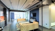 This single-floor space showcases a compelling blend of industrial and contemporary design. The open layout is defined by exposed ceiling ductwork and structural steel beams, which contrast elegantly with the clean white ceiling and polished dark hardwood flooring beneath. A striking navy blue accent wall serves as the focal point of the main lounge area, anchoring a sleek media console topped with a large flat-screen TV and tasteful décor accents. The seating arrangement features cream-toned tufted sofas and matching ottomans, lending a sophisticated yet comfortable atmosphere to the space. A vibrant patterned area rug adds a pop of color and warmth underfoot. Abundant natural light pours in through large glass doors at the far end of the space, opening the interior visually and creating an airy, welcoming feel. The overall ambiance balances the rawness of its industrial architectural elements — such as the visible HVAC components — with refined furnishings and thoughtful styling, making it well-suited for a variety of uses including lounging, entertaining, or collaborative gathering.