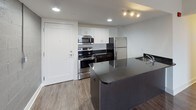 Located at 3075 W Grand Blvd in Detroit, Michigan, this single-floor unit offers a clean, contemporary design with functional layout and modern finishes throughout. Wood-look flooring runs seamlessly across the living, kitchen, and bedroom areas, lending warmth and visual continuity to the space.

The open-concept kitchen is well-appointed with stainless steel appliances — including a refrigerator, electric range, and over-the-range microwave — paired with white upper cabinetry, dark lower cabinetry, and a dark-toned island with an integrated sink. Recessed and track lighting illuminate the space effectively.

The living area is generously proportioned and filled with natural light from a large window offering views of the surrounding Detroit cityscape. An exposed, painted concrete block accent wall adds an industrial character to the space, complementing the otherwise neutral white walls.

A hallway connects the living area to the bedroom and bathroom. The bedroom is a well-sized room with a dedicated closet and its own accent of painted concrete block, maintaining the design continuity seen throughout the unit. The bathroom features white tile flooring, a bathtub, toilet, and recessed lighting with a grey accent wall. Additional in-unit utility access is conveniently located in the hallway area.