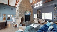 This clubhouse offers an impressive and welcoming gathering space defined by its soaring vaulted ceilings accented with rich, exposed wooden beams. At the heart of the main lounge stands a dramatic floor-to-ceiling stone fireplace, crafted from natural stacked stone and crowned with a mounted flat-screen television, serving as both a functional focal point and a striking architectural statement. The open layout of the lounge area is thoughtfully furnished with a curated mix of seating, including woven rattan accent chairs, upholstered armchairs, and plush sofas in neutral and navy tones, all arranged around a sleek metallic coffee table and a patterned blue area rug. Natural light streams in through multiple large windows positioned high along the walls, creating an airy and bright atmosphere throughout. A warm, ambient chandelier with an organic, branching design adds character overhead. Built-in shelving units along one wall provide both display space and visual depth to the room. The overall aesthetic masterfully combines rustic elements — exposed wood, natural stone, and warm textures — with refined, contemporary furnishings, resulting in a sophisticated yet comfortable communal environment ideal for socializing and relaxation.