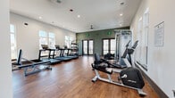 Located in Gallatin, Tennessee, this fitness center offers a comprehensive workout facility designed to accommodate a wide range of exercise routines. The space features warm hardwood-style flooring throughout, crisp white walls accented by a bold deep-green feature wall, and recessed lighting complemented by ceiling fans to maintain a comfortable environment during workouts.

Large windows line multiple walls, flooding the interior with abundant natural light and providing views of the surrounding community. Glass panel doors open directly to an outdoor area, enhancing the sense of openness and allowing for easy indoor-outdoor access.

The cardio zone is well-appointed with multiple treadmills and a variety of cycling and elliptical machines, offering ample options for aerobic training. A dedicated strength training area includes a multi-station cable machine, a full dumbbell rack, adjustable benches, and a functional training rig with TRX suspension straps. Additional equipment such as kettlebells, medicine balls, and a balance disc rounds out the functional fitness offerings. A wall-mounted television and a small storage unit with a mini-refrigerator provide added convenience. Floor-to-ceiling mirrors expand the sense of space while allowing users to monitor their form. The overall design balances functionality with a clean, modern aesthetic suited to an active lifestyle.