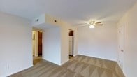 This apartment at The Devonshire Apartments is a single-story residence designed with a straightforward, functional layout. The main living area is finished with patterned wall-to-wall carpeting in neutral beige tones, complementing the clean white walls and white baseboards throughout. A ceiling fan with a built-in light fixture is centrally positioned in the living space, providing both air circulation and ambient lighting.

A structural half-wall divides the living area from the adjacent spaces, creating a subtle sense of separation while maintaining an open feel. Through the opening beside this divider, the kitchen is visible and features a white refrigerator and dark wood cabinetry, offering a warm contrast to the lighter tones of the living space. A hallway visible on the left side of the divider leads toward additional rooms, where dark wood doors and trim continue the cabinetry theme.

Overhead HVAC vents are integrated into the ceiling, ensuring climate control throughout the unit. The overall ambiance is clean, neutral, and move-in ready, with a simple palette that offers flexibility for personalization.