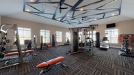 This fitness center is a comprehensively outfitted exercise facility designed to support a wide range of workout styles and fitness goals. The space is immediately distinguished by its striking geometric ceiling design, featuring an angular, faceted framework with recessed lighting and ceiling fans — creating an architectural focal point that elevates the overall aesthetic of the room.

The flooring throughout is a durable, patterned commercial carpet in dark tones of grey and charcoal, providing both comfort underfoot and a polished, modern look. Multiple large windows with blinds line the walls, inviting generous natural light and offering views of the surrounding landscape, contributing to an open and energizing atmosphere.

The strength training area is well-appointed with a multi-functional cable machine, a heavy punching bag, adjustable benches with orange upholstery, a weight rack with dumbbells, stability balls, resistance bands, and a plyo box. A dedicated stretching and flexibility zone features open floor space and storage shelving.

The cardio section includes treadmills, an elliptical, a rowing machine, and a stationary bike, all positioned near windows to make use of the natural views. Multiple wall-mounted flat-screen televisions are installed throughout the space for entertainment during workouts. A set of glass-paned double doors connects to an adjoining room, enhancing the facility's overall flow and accessibility.