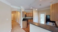 This single-floor residence offers a thoughtfully designed layout that balances functionality and comfort. The kitchen serves as a central highlight, featuring warm honey-toned wood cabinetry, dark granite-style countertops, and black appliances including a gas range and built-in microwave. A track lighting system overhead ensures excellent task illumination throughout the cooking space. The kitchen opens to a breakfast bar or peninsula, providing additional counter space and a casual dining option. An elegant arched entryway with a decorative column frames the transition between the kitchen and the adjoining living areas, lending a classic architectural touch to the open floor plan. Crown molding runs throughout, adding a refined finish to the ceilings. Hardwood flooring anchors the kitchen and entry areas, while plush carpeting extends into the living spaces for added warmth and comfort. A door with a sidelight window allows natural light to filter into the space, creating a bright and welcoming ambiance. The layout flows naturally between rooms, making the space feel open and connected. Overall, the residence combines traditional architectural details with practical design, creating a comfortable and inviting home environment.