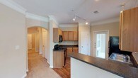 This single-floor residence offers a thoughtfully designed layout that balances functionality and comfort. The kitchen serves as a central highlight, featuring warm honey-toned wood cabinetry, dark granite-style countertops, and black appliances including a gas range and built-in microwave. A track lighting system overhead ensures excellent task illumination throughout the cooking space. The kitchen opens to a breakfast bar or peninsula, providing additional counter space and a casual dining option. An elegant arched entryway with a decorative column frames the transition between the kitchen and the adjoining living areas, lending a classic architectural touch to the open floor plan. Crown molding runs throughout, adding a refined finish to the ceilings. Hardwood flooring anchors the kitchen and entry areas, while plush carpeting extends into the living spaces for added warmth and comfort. A door with a sidelight window allows natural light to filter into the space, creating a bright and welcoming ambiance. The layout flows naturally between rooms, making the space feel open and connected. Overall, the residence combines traditional architectural details with practical design, creating a comfortable and inviting home environment.