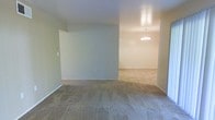 This single-floor residential space offers a straightforward and functional layout designed for comfortable everyday living. The interior is finished with wall-to-wall carpeting in a warm neutral tone that extends seamlessly throughout the main living areas, providing a cohesive and understated aesthetic. The walls are painted in soft, muted hues that lend a calm and welcoming ambiance to the space.

The main living area is open and unobstructed, offering a generous sense of space and flexibility for a variety of furniture arrangements. A partial wall creates a natural division between the living area and an adjoining room, which is visible in the background and features a classic chandelier suspended from the ceiling — a subtle decorative touch that adds a hint of traditional charm to the otherwise simple interior.

The layout flows naturally from one area to the next, making the space feel connected while still allowing for defined zones. Neutral-colored walls and consistent flooring tie the rooms together visually, while the clean lines and minimal architectural detailing give the space a blank-canvas quality, well-suited for personalization. Overall, this is a practical and move-in-ready single-floor residence with a calm, neutral palette and a functional open feel.