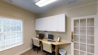 This single-floor business center offers a thoughtfully designed workspace tailored for productivity. The room features a long, built-in wooden workstation that comfortably accommodates two computer setups, each equipped with a desktop monitor, keyboard, and ergonomic cream-upholstered office chair. Above the desk, a row of white shaker-style wall cabinets with brushed nickel hardware spans the full width of the workspace, providing generous closed storage while maintaining a clean and organized appearance. The walls are finished in a soft, warm neutral tone, complemented by elegant crown molding that lends the space a polished, refined character. Overhead, a bright flush-mounted fluorescent light fixture ensures ample illumination throughout the room. A patterned area rug with warm earth tones adds a touch of comfort underfoot. Natural light filters in through a window on the adjacent wall, contributing to a bright and welcoming atmosphere. An open doorway leads to an adjoining area with additional shelving, enhancing the overall functionality of the space. This business center is well-suited for tasks requiring focus, connectivity, and convenient access to resources.