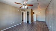 This single-floor space is characterized by dark hardwood-style flooring that runs consistently throughout the main living areas and bedrooms, lending a cohesive, grounded aesthetic. The main living area is outfitted with a ceiling fan with integrated lighting and opens directly toward the kitchen and bedroom access points, creating an efficient, open flow. Two closets are visible from the living area, offering generous built-in storage. A ceiling fan with a light fixture is present in each room, providing both comfort and illumination.

The kitchen is compact yet functional, featuring dark wood cabinetry, granite-style countertops, a mosaic tile backsplash, and white appliances including a refrigerator, electric range, and over-the-range microwave. A pendant light adds a subtle decorative touch above the kitchen area. One bedroom features a distinctive floor-to-ceiling wood-paneled accent wall, adding texture and character to the space. Another bedroom includes a window dressed with red curtains, allowing natural light to filter in.

The bathroom is well-appointed with a dark wood vanity, mosaic tile countertop, tiled walls, a window for natural light, and a full soaking tub, all set atop durable tile flooring.