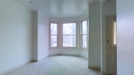 This single-floor space is defined by a standout curved bay window alcove, which serves as the room's focal point. The bay consists of three double-hung windows with warm wood framing, arranged in a gentle arc that floods the interior with natural light and frames views of mature trees and greenery outside. The soft, diffused daylight creates a bright and airy atmosphere throughout the room.

The walls are finished in a clean, neutral white, complemented by subtle crown molding that adds a touch of architectural refinement. Plush, light-toned carpeting covers the floor, contributing to the room's calm and understated character. A floor vent is positioned beneath the bay window, indicating in-floor climate control.

The layout is open and versatile, offering a blank canvas well-suited for a variety of uses. The curved bay alcove provides an inviting nook ideal for seating or reading. The overall ambiance is quiet and understated, with the natural light and neutral palette creating a welcoming, move-in-ready environment that balances simplicity with classic architectural detail.