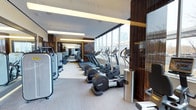 This professionally designed fitness center offers a comprehensive workout environment within a single, expansive floor. The space is characterized by its sleek, contemporary aesthetic, combining rich dark wood wall paneling with a neutral light-toned flooring to create a refined yet functional atmosphere. Floor-to-ceiling windows line one side of the facility, flooding the interior with abundant natural light and providing open views of the surrounding cityscape — an inviting backdrop for any workout. The cardio zone is prominently featured along the window-facing wall and is outfitted with a full row of treadmills, elliptical trainers, and other cardio machines, all equipped with individual digital displays. Strength and resistance training equipment is organized along the opposite side, offering a wide variety of machines to support a full-body training regimen. Recessed and linear pendant lighting ensures bright, even illumination throughout. Reflective surfaces and mirrors enhance the sense of openness and allow users to monitor their form. The overall layout is thoughtfully organized to allow for smooth traffic flow between equipment zones, making this fitness center a well-appointed and functional space suited for a wide range of training needs.