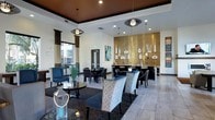 This single-floor communal space exudes a refined, contemporary aesthetic designed for comfort and social interaction. The layout is open and expansive, seamlessly blending multiple seating arrangements that invite conversation and relaxation. A dramatic gold mosaic tile accent wall serves as the room's centerpiece, drawing the eye immediately and anchoring a bar or service counter area beneath it. Decorative shelving mounted on the gold wall displays artistic vases and accents, adding a curated, gallery-like touch.

The space features a rich mix of seating options, including patterned upholstered chairs, sleek dark accent chairs in deep teal and charcoal, and polished dark tables that contrast elegantly against the light tile flooring. A tray ceiling with a warm wood finish adds architectural depth overhead, complemented by a series of distinctive oval-shaped pendant lights that provide ambient illumination alongside recessed lighting throughout.

Floor-to-ceiling windows dressed with layered brown drapery allow natural light to filter in while maintaining a sense of privacy. Artwork adorns the crisp white walls, contributing to the polished, upscale atmosphere. Overall, the space presents a well-appointed, modern lounge environment.