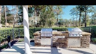 This outdoor amenity space is designed to offer residents a comprehensive recreational experience in a welcoming, resort-inspired setting. At the heart of the area is a generously sized swimming pool with a smooth concrete surround, lined with lounge chairs perfect for sunbathing. A classic white-columned pergola provides shaded seating with glass-top tables and vibrant teal chairs accented by bold red umbrellas, creating a comfortable outdoor dining atmosphere. The standout feature of this space is the built-in outdoor kitchen, constructed with stacked natural stone bases and outfitted with multiple stainless steel grills — designated for pool guests — making it ideal for gatherings and cookouts. Beyond the wrought iron perimeter fencing, lush landscaping and mature trees frame the area beautifully, with a serene water feature visible in the background, adding to the tranquil ambiance. The multi-story residential buildings surrounding the amenity area provide a sense of community and close convenience. The entire space benefits from abundant natural light and open sky views, creating an inviting environment year-round for socializing, relaxing, and enjoying outdoor living.