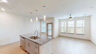 Located at 8025 Monty Stratton Pkwy in Greenville, Texas, this thoughtfully designed single-story residence offers a clean, modern aesthetic throughout its efficiently laid-out floor plan. Light wood-style flooring flows through the main living areas, complementing the bright, neutral color palette that gives the space an open and airy feel.

The open-concept kitchen and living room serve as the heart of the home. The kitchen is outfitted with gray shaker-style cabinetry, stainless steel appliances including a refrigerator, range, and built-in microwave, and a central island topped with granite countertops. Stylish pendant lighting hangs above the island, adding a modern touch. The layout allows for seamless interaction between the kitchen and living areas.

The bedroom features plush carpet flooring, a ceiling fan, and multiple windows dressed with blinds that invite natural light and outdoor views of the surrounding greenery. A dedicated walk-in closet with built-in shelving and hanging rods provides ample storage. A separate laundry room accommodates a full-size washer and dryer.

The bathroom is finished with a granite-topped vanity with gray cabinetry, light wood-look tile flooring, and a soaking tub with a tiled surround and sliding glass enclosure, combining both comfort and style.