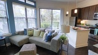 This single-floor residence offers an open-concept layout that integrates the living area and kitchen into one cohesive, functional space. The living room is anchored by a generously sized sectional sofa, complemented by dark hardwood flooring that runs throughout, adding warmth and continuity to the interior. Large floor-to-ceiling windows and a sliding glass door flood the space with natural light and frame views of lush greenery beyond the private balcony, creating a seamless indoor-outdoor connection. The balcony, accessible directly from the living area, provides an intimate outdoor retreat overlooking mature trees and neighboring landscaping.

The kitchen sits adjacent to the living space, separated by a raised countertop bar that doubles as a casual dining surface. Dark cabinetry contrasts with light countertops, and stainless steel appliances, including a full-sized refrigerator, contribute a modern, polished aesthetic. Pendant lighting above the kitchen area adds a refined decorative touch alongside a wall-mounted sconce. The overall ambiance is bright, contemporary, and inviting, with thoughtful design details that balance style and everyday livability.