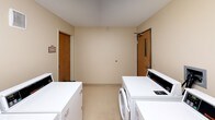 This shared laundry facility is a well-organized, single-floor space designed to accommodate the laundering needs of multiple users efficiently. The room is finished with neutral beige walls and light tile flooring, creating a clean and functional environment. Multiple commercial-grade, top-loading washing machines and dryers are arranged along the walls, providing ample capacity for simultaneous use. Each machine features coin-operated controls, and laundry tip signage is posted on the wall for user guidance. Overhead fluorescent lighting ensures the space is well-lit throughout, while a ventilation grille on the upper wall supports proper airflow. The room benefits from two access doors — one serving as the main entry and another leading to a designated storage room, clearly marked with signage. The storage room offers additional utility for the facility. The overall layout is straightforward and practical, maximizing the usable floor space while keeping equipment accessible. The textured ceiling and simple architectural design reflect a no-frills, utilitarian approach focused entirely on functionality and ease of use.
