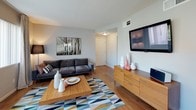 This ground-floor unit offers a well-organized single-level layout with two bedrooms and one bathroom. The living room is a bright, welcoming space featuring hardwood-style flooring, neutral walls, and a sliding glass door fitted with vertical blinds that opens directly onto a private patio, allowing natural light and outdoor views to enhance the interior. A wall-mounted television and a warm wood media console complement the room's contemporary aesthetic.

Adjacent to the living area, the dining space is framed by large windows with floor-length draping curtains, providing ample natural light and views of the surrounding exterior. The kitchen features white cabinetry, granite-style countertops, a stainless steel dishwasher, and a window above the sink.

The primary bedroom features a bold crimson accent wall, carpeted flooring, a built-in closet, and a natural light source through a window with draping curtains. The second bedroom also offers carpeted flooring, a soft blue accent wall, and mirrored sliding closet doors. The bathroom includes a tub-shower combination with white tile surround, white cabinetry, and hardwood-style flooring.