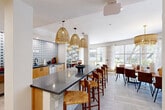 Located at 4111 N Drinkwater Blvd in Scottsdale, Arizona, this thoughtfully designed kitchen and dining space offers a cohesive blend of modern and bohemian aesthetics across a single open floor. The kitchen is anchored by warm natural wood cabinetry paired with sleek dark countertops, including a generously sized island that serves as both a workspace and a casual gathering spot with woven rush-seat bar stools. A striking geometric patterned tile backsplash in blue and white adds a bold visual accent, while a series of oversized woven pendant lights hang above the island, lending warmth and texture to the space. The kitchen is fully equipped with a sink, appliances, and ample storage along both sides of the galley-style run. The adjacent dining area features a substantial live-edge wood table surrounded by leather upholstered chairs, crowned by an elegant gold fringe chandelier. Floor-to-ceiling windows wrap around the dining and lounge areas, flooding the space with natural light and offering panoramic views of mature palm trees and lush desert landscaping. A cozy lounge nook with rattan chairs and a low round coffee table sits just beside the windows, creating an inviting retreat. Light-toned large-format tile flooring unifies the entire space, enhancing its bright, airy ambiance.