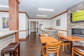 This single-story space is designed as a welcoming communal gathering area, combining dining, lounge, and service functions within an open, cohesive layout. The room is anchored by a striking stacked-stone fireplace wall, which also hosts a wall-mounted flat-screen television, creating a natural focal point for the dining area. Multiple wooden tables with ladder-back chairs are arranged throughout, providing ample seating for guests.

A built-in service counter runs along one side of the room, featuring warm wood cabinetry, a stone veneer base, and a laminate countertop. A raised display ledge above the counter is accented with decorative planters, adding a refined touch. Structural columns with matching stone and wood detailing define zones within the open floor plan without disrupting the flow of the space.

A separate nook off the main area offers a quieter retreat, furnished with a cushioned armchair and a modern open shelving unit. Natural light filters in through blinds-covered windows, complemented by recessed fluorescent ceiling fixtures. Dark hardwood flooring, rich wood crown molding, and neutral walls unify the space, giving it a comfortable, polished ambiance suited for relaxed social gatherings.