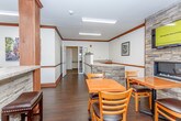 This single-story space is designed as a welcoming communal gathering area, combining dining, lounge, and service functions within an open, cohesive layout. The room is anchored by a striking stacked-stone fireplace wall, which also hosts a wall-mounted flat-screen television, creating a natural focal point for the dining area. Multiple wooden tables with ladder-back chairs are arranged throughout, providing ample seating for guests.

A built-in service counter runs along one side of the room, featuring warm wood cabinetry, a stone veneer base, and a laminate countertop. A raised display ledge above the counter is accented with decorative planters, adding a refined touch. Structural columns with matching stone and wood detailing define zones within the open floor plan without disrupting the flow of the space.

A separate nook off the main area offers a quieter retreat, furnished with a cushioned armchair and a modern open shelving unit. Natural light filters in through blinds-covered windows, complemented by recessed fluorescent ceiling fixtures. Dark hardwood flooring, rich wood crown molding, and neutral walls unify the space, giving it a comfortable, polished ambiance suited for relaxed social gatherings.