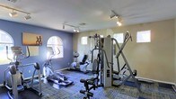 This fitness center offers a comprehensive workout environment designed to accommodate a range of exercise needs. The space features a diverse selection of equipment, including an elliptical trainer, a stationary recumbent bike, a rowing machine, and a multi-station cable weight machine, providing options for both cardiovascular and strength training workouts. An additional stair climber rounds out the cardio offerings. The room is finished with patterned, blue-toned flooring that adds a distinctive visual character to the space. Walls are painted in a two-tone scheme, with a deep blue accent wall on one side and neutral beige tones on the others, creating a motivating and dynamic atmosphere. Natural light filters into the room through an arched window and several smaller rectangular windows positioned along the upper walls, keeping the space bright and airy. Overhead track lighting supplements the natural light, ensuring even illumination throughout the facility. The ceiling features a standard finish with visible ventilation, maintaining comfort during workouts. Overall, the fitness center is thoughtfully arranged to maximize usability while maintaining an open, energetic feel.