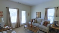 This single-floor space offers a warm and inviting atmosphere with a thoughtfully designed layout. The living area serves as the heart of the home, bathed in natural light streaming through large windows dressed with elegant grommet-style curtains in soft neutral tones. A glass-paneled door provides direct access to the outdoors, further enhancing the sense of openness and connectivity to the exterior.

The living room features plush, light-toned carpeting underfoot, complementing the neutral wall palette that creates a calm and airy ambiance. A generous sofa, accented with warm rust and patterned throw pillows, anchors the seating area, while a classic wooden coffee table with decorative accessories adds a touch of refined character. Framed botanical wall art ties the earthy color palette together, giving the space a cohesive and welcoming feel.

Ceiling-mounted air vents suggest a central climate control system, contributing to year-round comfort. The overall design balances functionality with aesthetic appeal, making it a practical yet stylish living environment. The single-floor layout ensures easy accessibility throughout, and the abundant natural light lends the space an uplifting, open quality that is both visually pleasing and livable.