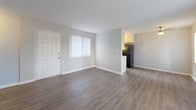 This single-floor residence offers a well-organized layout designed for comfortable everyday living. The open living area greets you at the entrance, featuring durable wood-look laminate flooring, neutral gray walls, and clean white baseboards that create a bright, modern aesthetic. Vertical blinds on the windows allow natural light to fill the space, enhancing the airy, open feel of the main living zone.

The galley-style kitchen is efficiently designed, running along both walls to maximize functionality. It features white shaker-style cabinetry with brushed nickel hardware, white countertops, a stainless steel double-basin sink, and black appliances including a full-size refrigerator. A window at the end of the kitchen brings in additional natural light and ventilation. Exposed brick accents on one wall add a subtle textural contrast to the otherwise clean, contemporary finishes.

The bedrooms are carpeted for comfort and finished in the same soft neutral palette. Each room benefits from windows dressed with vertical blinds, providing ample natural light and views of the surrounding greenery. One bedroom includes a closet with bi-fold doors, offering practical storage. HVAC vents are present throughout, ensuring climate control across the entire floor plan. Overall, the space presents a clean, move-in-ready environment with consistent, modern finishes.