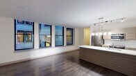 This contemporary single-floor residence showcases a thoughtfully designed open-concept layout that seamlessly blends living and kitchen spaces. Rich, dark hardwood flooring extends throughout the main living areas, creating a warm contrast against the crisp white ceilings and neutral walls. A row of large, floor-to-near-ceiling windows lines the living area, flooding the space with natural light and framing views of the surrounding urban streetscape.

The kitchen is a standout feature, centered around a generous island with a clean white countertop and a wood-toned base that mirrors the cabinetry. Stainless steel appliances — including a full-size refrigerator, range, and microwave — are neatly integrated alongside the cabinetry. A modern pendant light fixture with multiple hanging glass shades illuminates the island, complemented by a track lighting system overhead.

A separate bedroom offers a quieter retreat, finished with plush carpeting and two windows that bring in additional natural light and city views. The bathroom is finished with light tile flooring, a combination tub and shower with clean white tile surround, and a wood-accented vanity with a light countertop. Throughout, the design maintains a cohesive modern aesthetic with clean lines, neutral tones, and quality finishes.