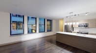 This contemporary single-floor residence showcases a thoughtfully designed open-concept layout that seamlessly blends living and kitchen spaces. Rich, dark hardwood flooring extends throughout the main living areas, creating a warm contrast against the crisp white ceilings and neutral walls. A row of large, floor-to-near-ceiling windows lines the living area, flooding the space with natural light and framing views of the surrounding urban streetscape.

The kitchen is a standout feature, centered around a generous island with a clean white countertop and a wood-toned base that mirrors the cabinetry. Stainless steel appliances — including a full-size refrigerator, range, and microwave — are neatly integrated alongside the cabinetry. A modern pendant light fixture with multiple hanging glass shades illuminates the island, complemented by a track lighting system overhead.

A separate bedroom offers a quieter retreat, finished with plush carpeting and two windows that bring in additional natural light and city views. The bathroom is finished with light tile flooring, a combination tub and shower with clean white tile surround, and a wood-accented vanity with a light countertop. Throughout, the design maintains a cohesive modern aesthetic with clean lines, neutral tones, and quality finishes.