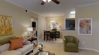 This single-floor residence offers a thoughtfully designed open-concept layout that connects the living, dining, and kitchen areas into one cohesive, flowing space. The living area is welcoming and comfortable, highlighted by crown molding, a ceiling fan, and warm neutral wall tones that create a relaxed ambiance. Plush carpeting runs throughout the main living spaces, adding softness underfoot. A decorative chandelier anchors the dining area, where a round sunburst mirror serves as a striking focal point on the wall. The dining space comfortably accommodates a full table and chair set, ideal for everyday meals or entertaining. The kitchen is accessible via a raised breakfast bar with granite-style countertops, providing both a casual dining option and a natural visual divider between the kitchen and living areas. Dark cabinetry and built-in appliances, including a microwave and oven, give the kitchen a sleek, modern feel. A hallway visible from the living room leads to additional private areas of the residence. Natural and ambient lighting work together to keep the interior bright and inviting. Overall, the space presents a balanced combination of functionality and style, making it a comfortable and well-organized home.