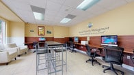 This single-floor United Property Management office is designed to efficiently serve clients and staff in a welcoming, well-organized environment. The space is divided into several functional zones, each thoughtfully appointed to support day-to-day operations.

The largest area features an extended built-in workstation along the perimeter wall, finished in warm cherry-toned wood paneling that runs as a wainscot throughout the room. Multiple desktop computers are arranged along this counter, each accompanied by ergonomic mesh office chairs, providing ample workspace for staff or client use. Above the workstation area, the United Property Management branding and tagline — "Paying attention to every detail" — is prominently displayed on the soft yellow walls.

A central high-top table with bar-height stools offers additional flexible workspace. Toward the front of the office, a welcoming lounge area features cream leather seating arranged near large windows, which frame views of the surrounding streetscape and allow generous natural light to fill the room.

A utility area near the rear accommodates a multifunction copier and additional storage cabinetry, alongside an interior window that connects to a back office or corridor. Light tile flooring and a drop-panel ceiling with flush lighting tie the entire space together, creating a clean, professional ambiance throughout.